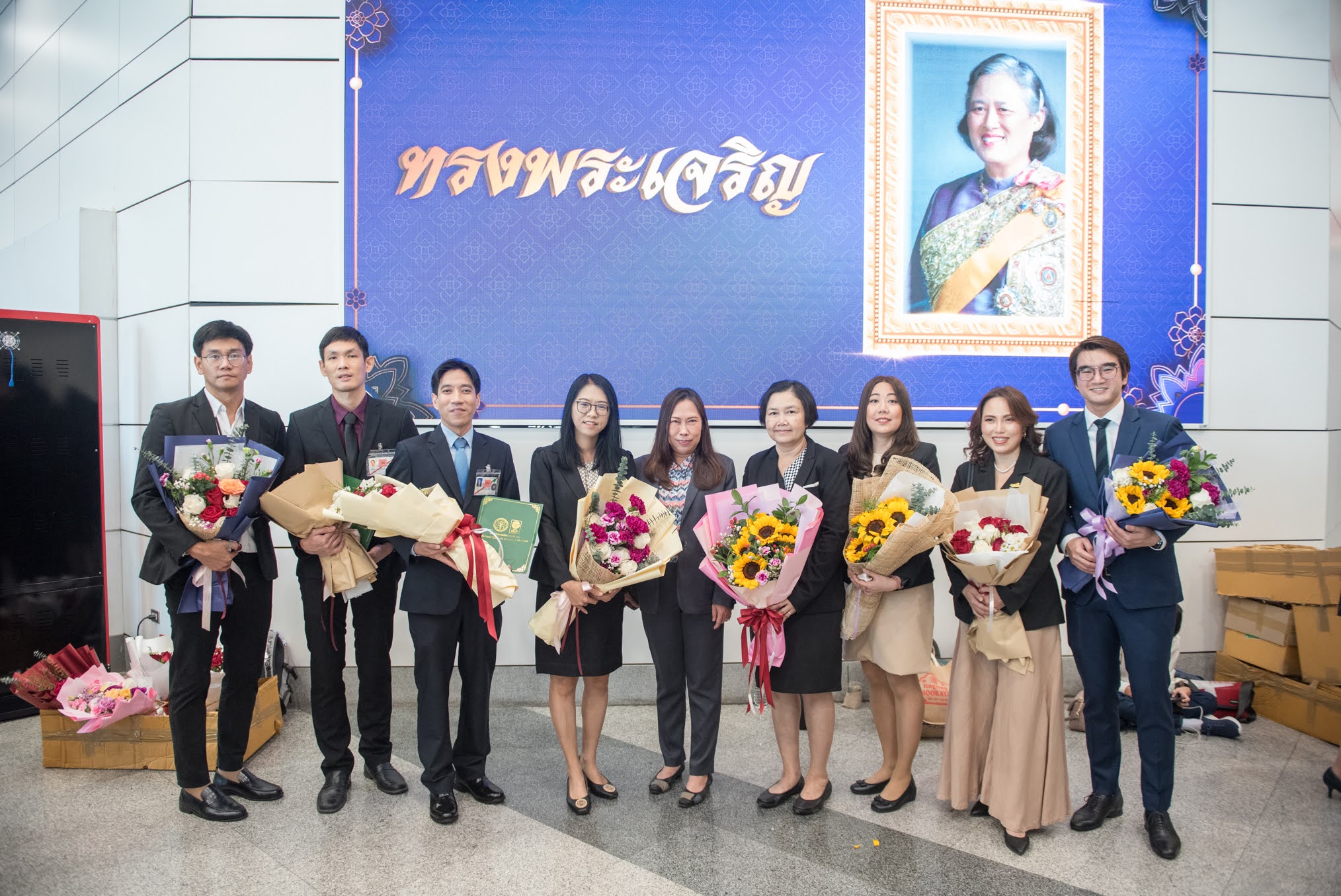 ภาพถ่ายกลุ่มจำนวน 9 ท่านในงาน “วันนักประดิษฐ์ ประจำปี 2568” โดยกลุ่มคนจำนวนมากยืนแพวกเขาสวมชุดสูทและถือช่อดอกไม้หลากสี มีพื้นหลังเป็นป้ายสีน้ำเงินแสดงคำว่า ทรงพระเจริญ