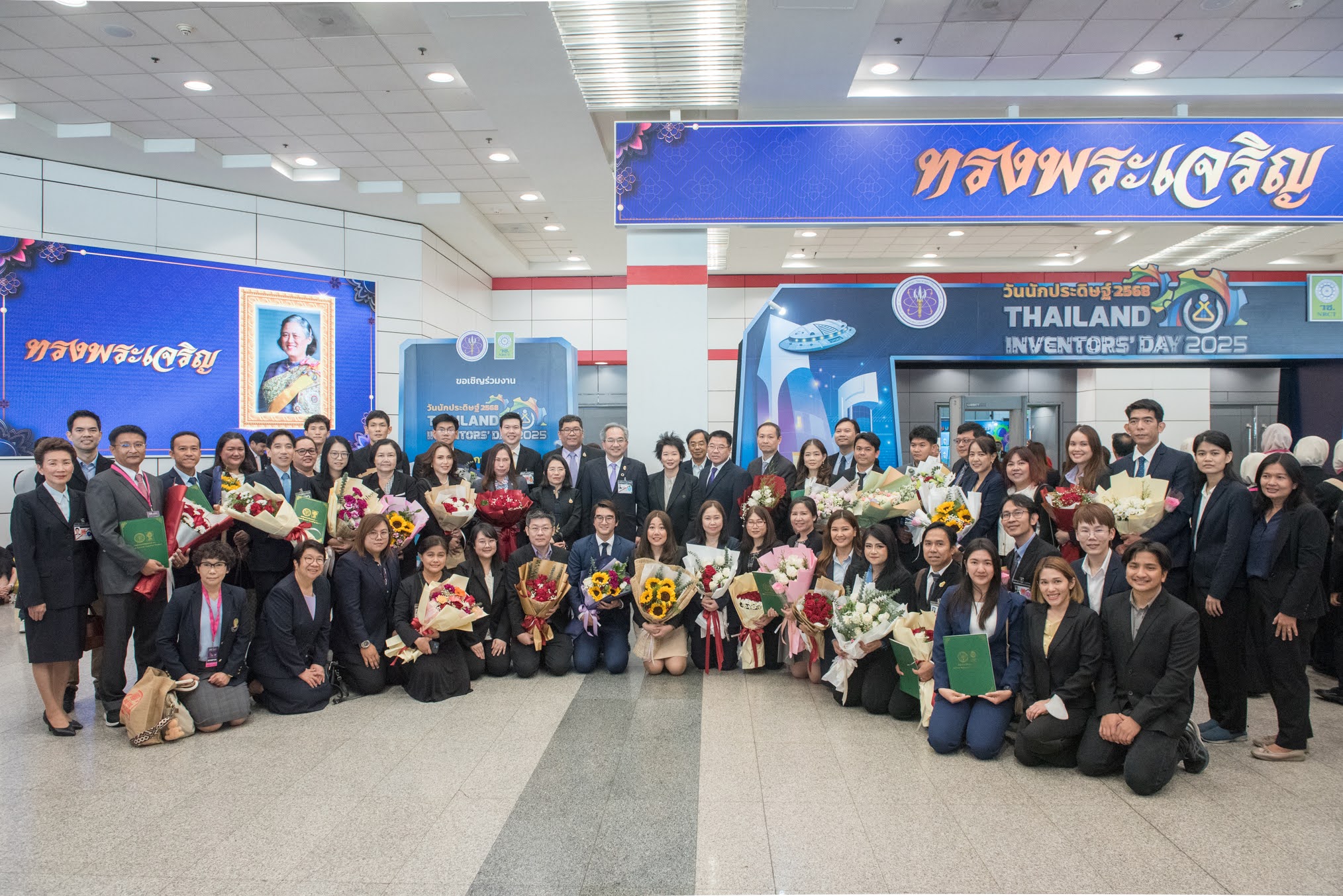 ภาพถ่ายกลุ่มคนในงาน “วันนักประดิษฐ์ ประจำปี 2568” โดยกลุ่มคนจำนวนมากยืนและนั่งรวมกันในห้องโถงพวกเขาสวมชุดสูทและถือช่อดอกไม้หลากสี มีพื้นหลังเป็นป้ายสีน้ำเงิน 