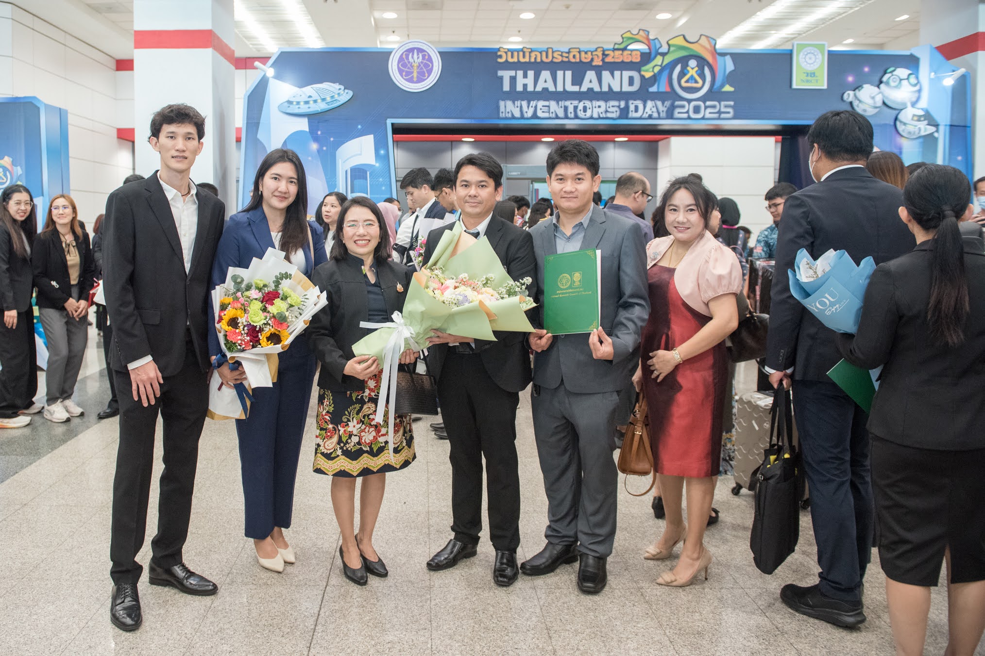 ภาพถ่ายกลุ่มจำนวน 5 ท่านในงาน “วันนักประดิษฐ์ ประจำปี 2568” โดยกลุ่มคนจำนวนมากยืนแพวกเขาสวมชุดสูทและถือช่อดอกไม้หลากสี มีพื้นหลังเป็นป้ายสีน้ำเงินแสดงคำว่า วันนักประดิษฐ์ 2568 และ Thailand invertors'day 2025