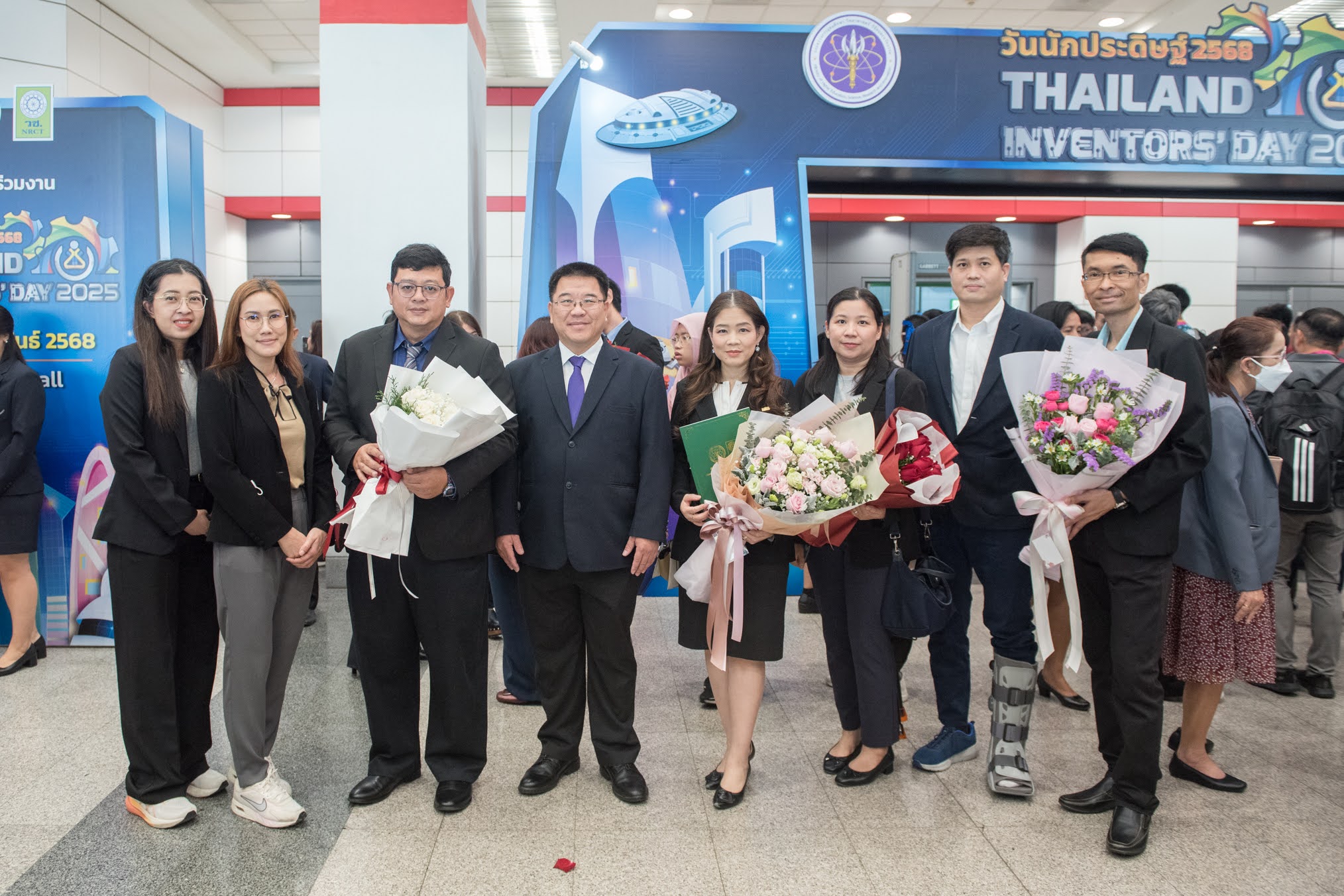 ภาพถ่ายกลุ่มจำนวน 7 ท่านในงาน “วันนักประดิษฐ์ ประจำปี 2568” โดยกลุ่มคนจำนวนมากยืนแพวกเขาสวมชุดสูทและถือช่อดอกไม้หลากสี มีพื้นหลังเป็นป้ายสีน้ำเงินแสดงคำว่า วันนักประดิษฐ์ 2568 และ Thailand invertors'day 2025