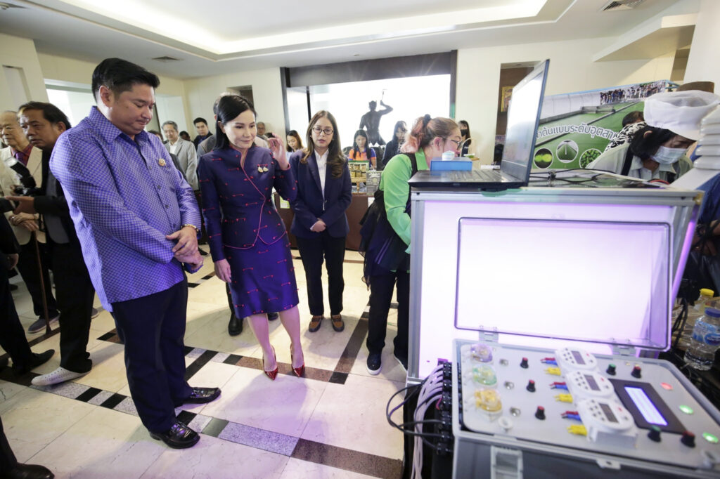 ภาพนี้แสดงให้เห็นกลุ่มคนที่กำลังเยี่ยมชมงานนิทรรศการหรือการนำเสนอเทคโนโลยีบางอย่างภายในอาคารที่มีบรรยากาศเป็นทางการ บุคคลสำคัญในภาพ ได้แก่ หญิงสาวที่แต่งชุดสูทเดรสสีน้ำเงินเข้มลายแดง คือศาสตราจารย์ ดร.นฤมล ภิญโญสินวัฒน์ รัฐมนตรีว่าการกระทรวงเกษตรและสหกรณ์ และชายในเสื้อเชิ้ตลายสีน้ำเงิน ดร.ศุภนิจ พรธีระภัทร นักวิจัยอาวุโส กำลังให้ความสนใจกับอุปกรณ์เทคโนโลยีที่มีไฟเรืองแสงและหน้าจอแสดงผล ขณะที่ผู้ร่วมงานคนอื่นๆ กำลังสังเกตการณ์และอธิบายข้อมูลเกี่ยวกับอุปกรณ์ดังกล่าว ในพื้นหลัง มีป้ายประชาสัมพันธ์เกี่ยวกับโครงการที่ดูเหมือนจะเกี่ยวข้องกับเทคโนโลยีด้านเกษตรกรรมหรือสิ่งแวดล้อม โดยมีภาพถ่ายและข้อมูลแสดงอยู่บนแบนเนอร์ รวมถึงผู้เข้าร่วมงานที่สวมชุดป้องกันหรือหน้ากากอนามัย ซึ่งอาจบ่งบอกถึงมาตรการด้านสุขอนามัยภายในงาน