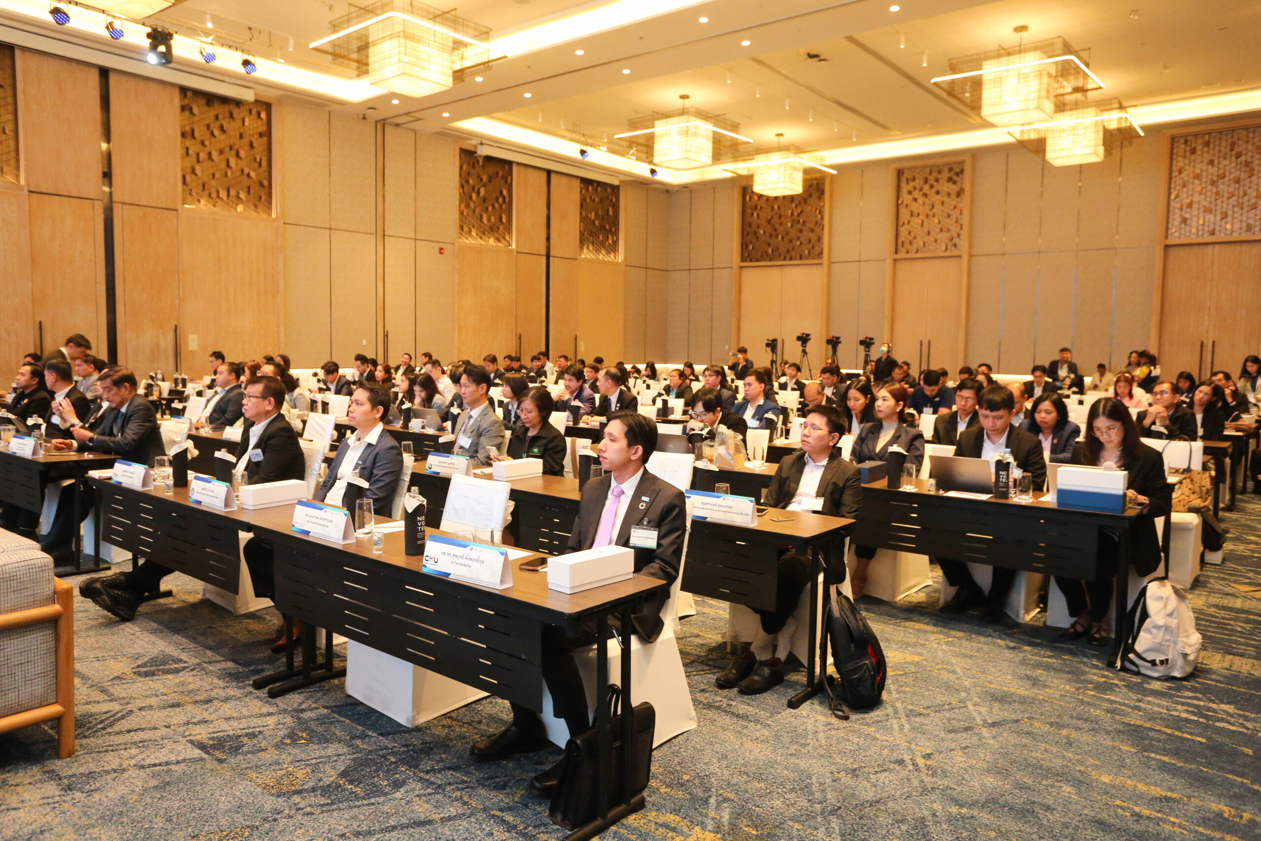 ภาพบรรยากาศการประชุมในห้องประชุมโรงแรม ผู้เข้าร่วมจำนวนมากนั่งที่โต๊ะประชุม พร้อมป้ายชื่อ อุปกรณ์ประชุม และกล่องของว่าง