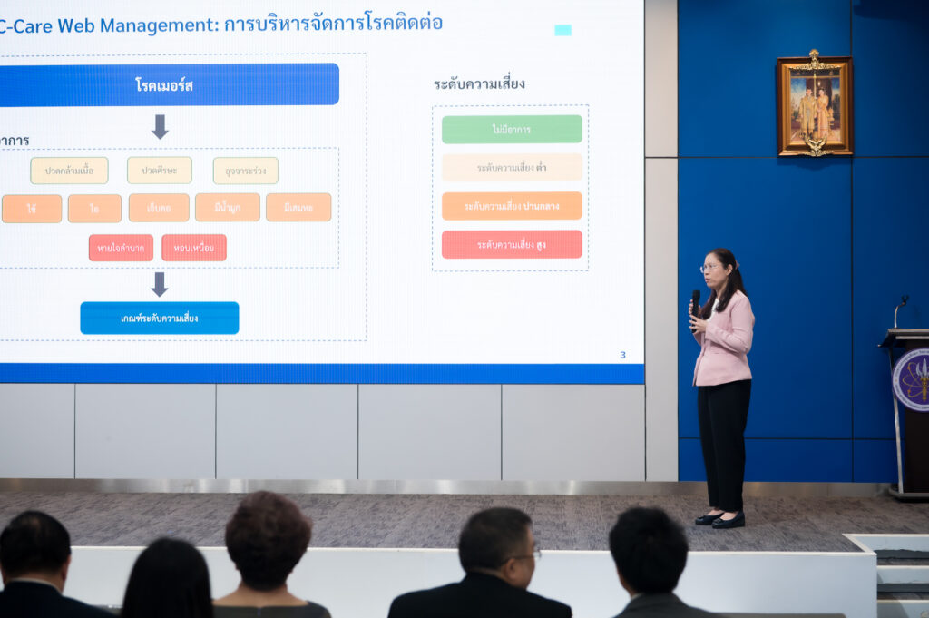 ภาพ ดร.อนันต์ลดา โชติมงคล กำลังยืนบรรยายเนื้อหา DDC-Care Platform ระบบเฝ้าระวังโรคติดต่อ ควบคู่กับสไลด์ที่เปิดบนจอ LED บนเวที