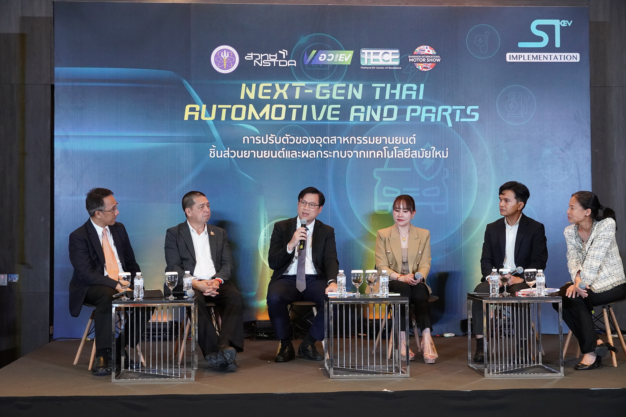 ภาพ ผู้เชี่ยวชาญในแวดวงอุตสาหกรรม นั่งเก้าอี้คุยสัมมนากัน โดยมีบางท่านถือไมค์กำลังพูดแลกเปลี่ยนความคิด หน้า Backdrop บนเวที 