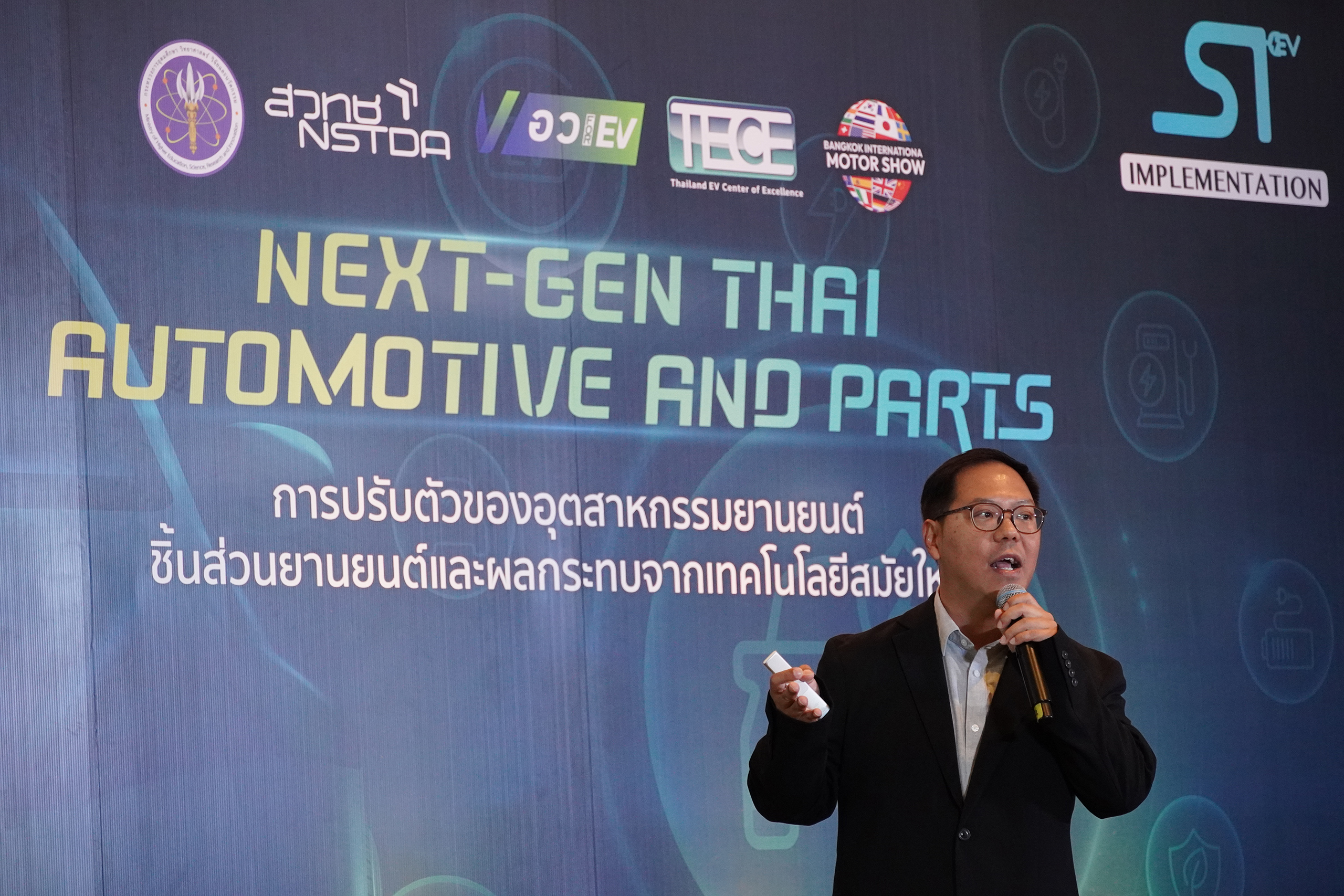 คุณปิยพงศ์ เปรมวรานนท์ หัวหน้าทีมวิจัย เอ็มเทค  ยืนถือไมค์หน้า Backdrop บนเวที กำลังนำเสนอผลงาน