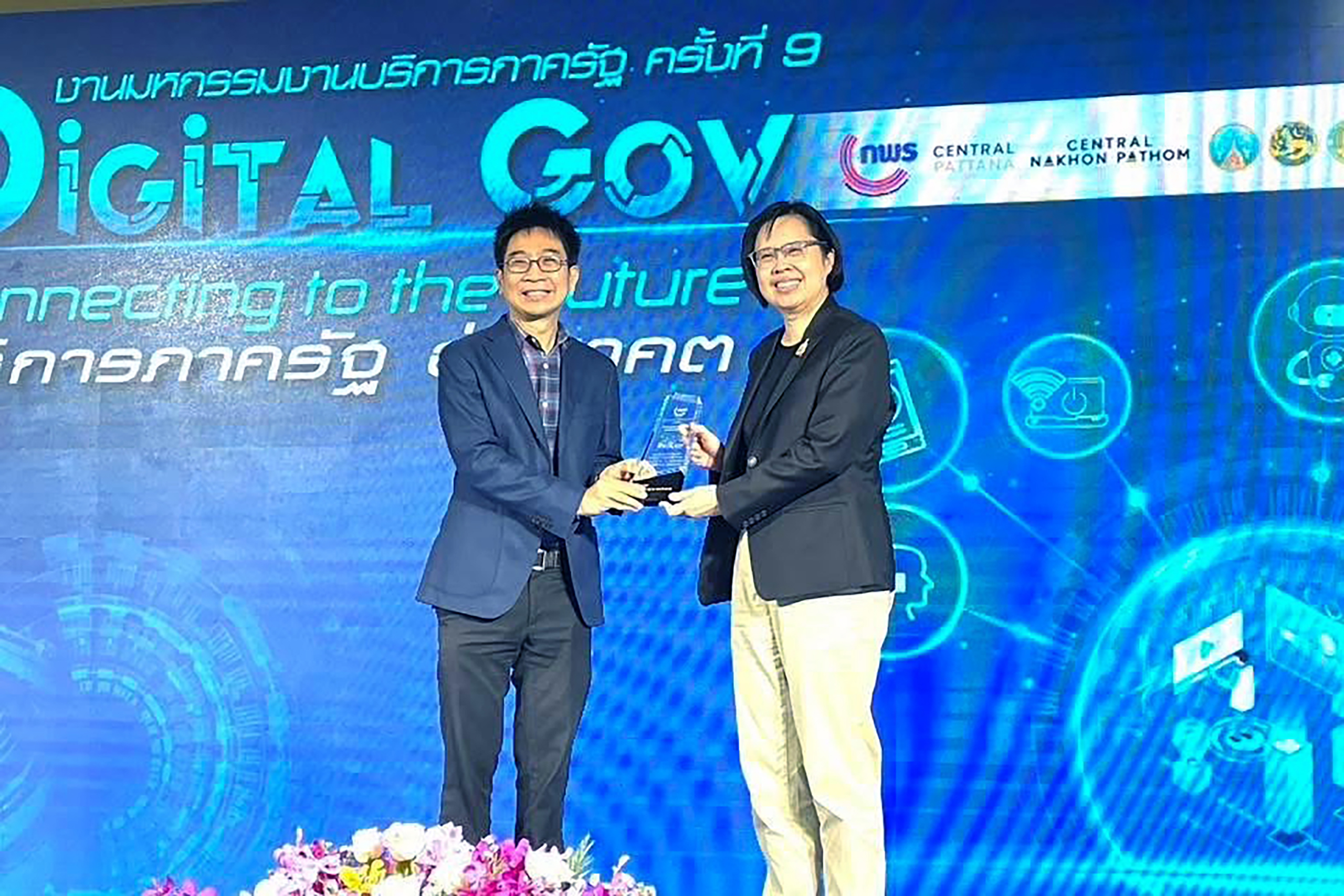 ภาพ ดร.วสันต์ ภัทรอธิคม ผู้ช่วยผู้อำนวยการ สวทช. ยืนรับโล่จาก น.ส.อ้อนฟ้า เวชชาชีวะ เลขาธิการ ก.พ.ร. ซึ่งเป็นประธานจัดงาน ด้านหลังเป็น blackdrop งาน ตรีมสีน้ำเงิน
