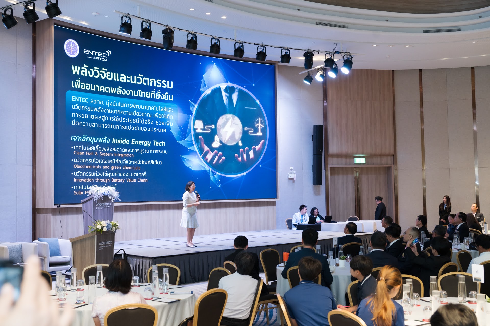 ภาพเต็มตัว ของ ดร.สุมิตรา กำลังถือไมค์บรรยาย บนเวที ด้วยชุดสีขาวครีม ด้านหลังเป็น ข้อมูลที่ขึ้น จอ LED ด้านเป็นผู้เข้าร่วมสัมมนา