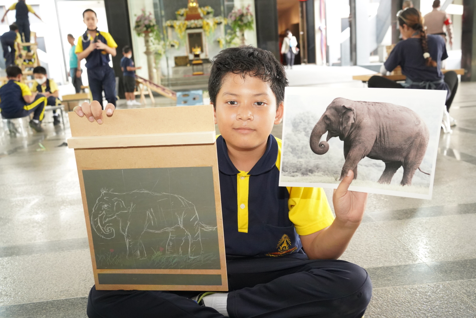 เด็กชายสวมชุดนักเรียนถือผลงานภาพวาดช้างที่วาดบนกระดานพาราโน้ต 