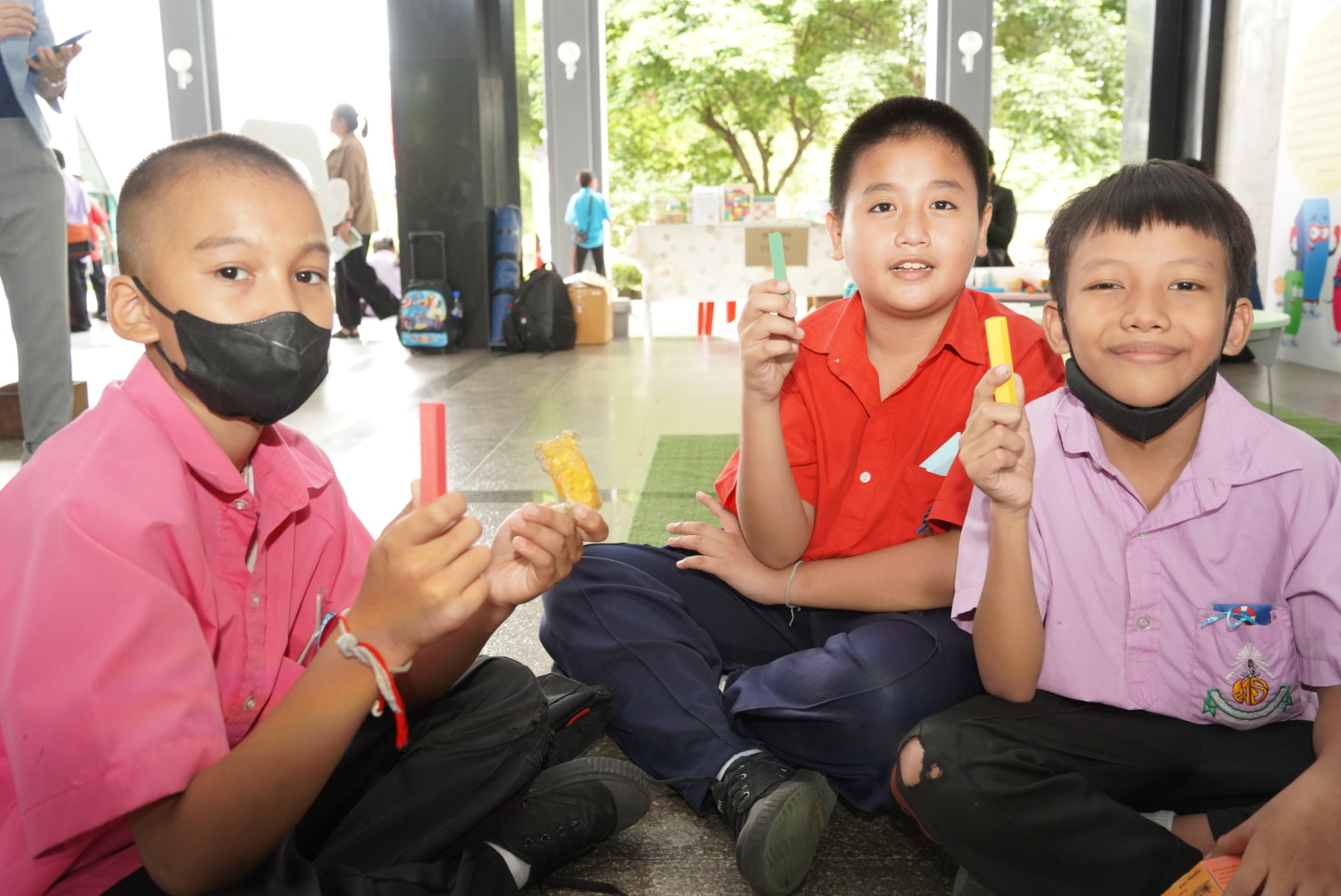 เด็กชายสามคนในชุดนักเรียนต่างโรงเรียน ยิ้มถือแท่งพาราโน้ตสีต่าง ๆ โชว์ผลงานกิจกรรมพร้อมรอยยิ้มสนุกสนาน