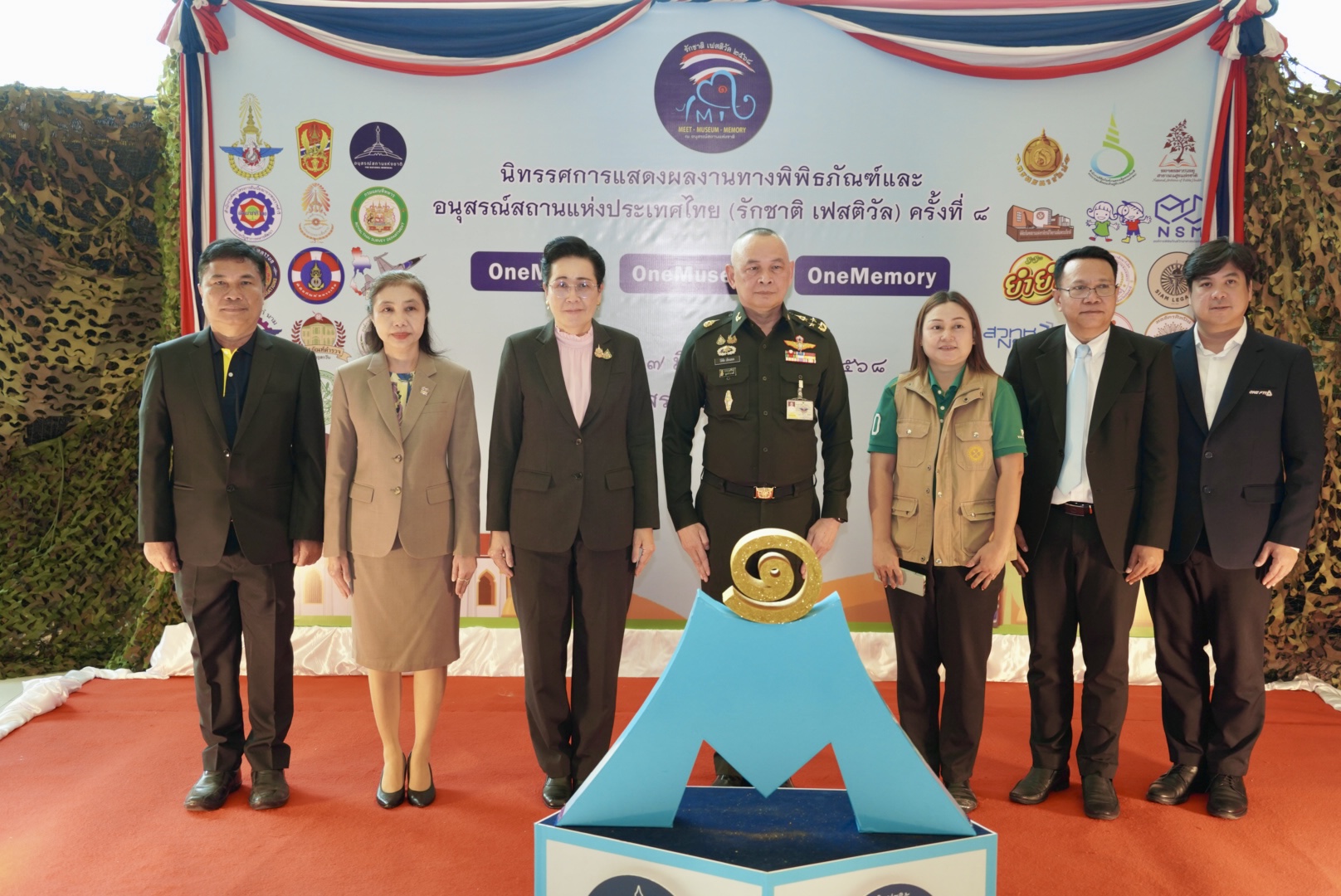 ภาพผู้บริหารและผู้แทนจากหน่วยงานต่าง ๆ รวม 8 คน ยืนถ่ายภาพร่วมกันบนเวทีในพิธีเปิดงาน "รักชาติ Festival ครั้งที่ 8" ณ อนุสรณ์สถานแห่งชาติ โดยมีฉากหลังเป็นป้ายโลโก้หน่วยงานที่เข้าร่วมจัดนิทรรศการ
