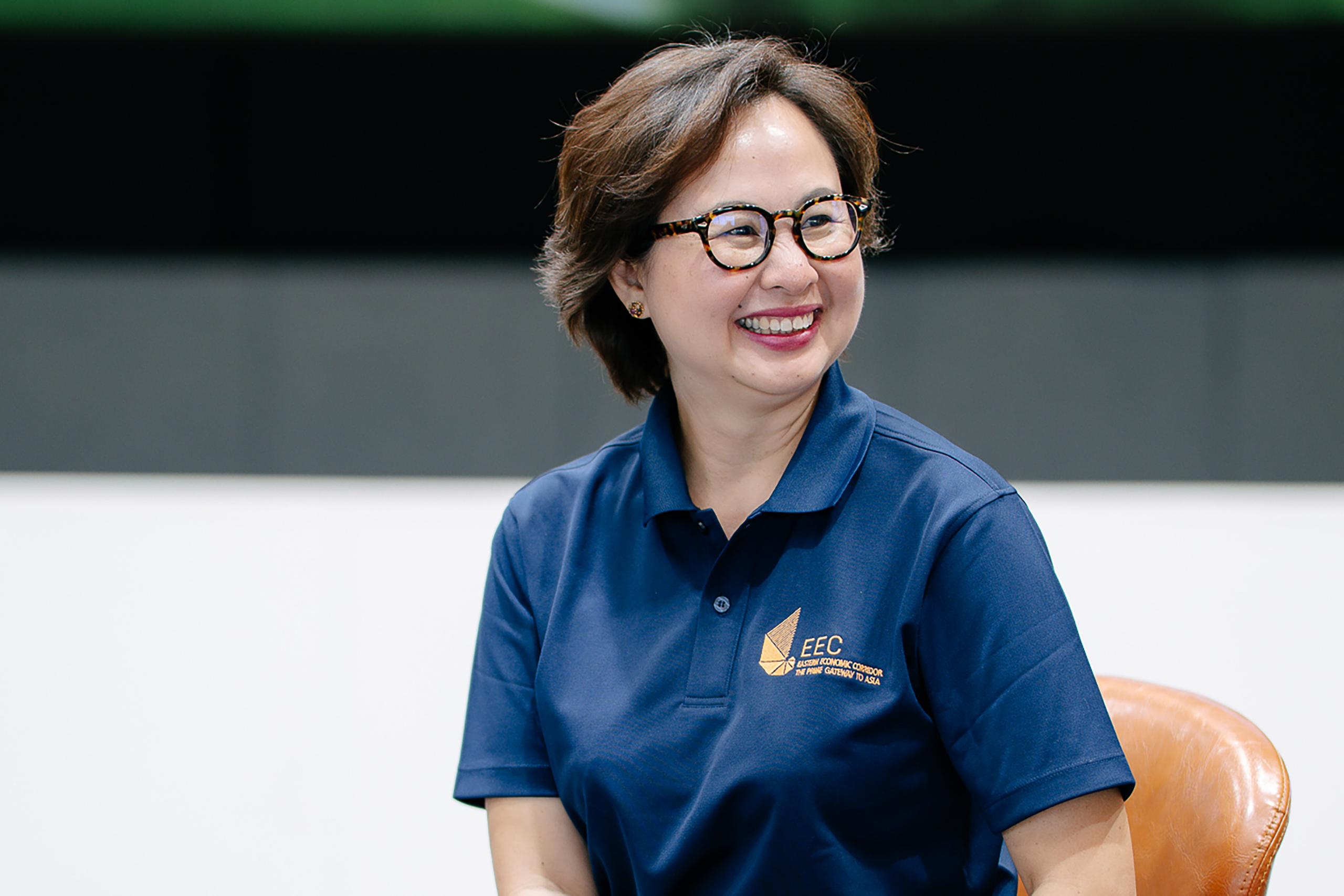 ภาพครึ่งตัว ดร.จุฑา นั่งเก้าอี้ พร้อมใบหน้ากำลังยิ้มเห็นฟัน ใส่แว่นสายตา ผมสีน้ำตาลแดง สวมเสื้อโปโลกรมท่า โลโก้ EECi ด้านหน้าอกข้างซ้าย