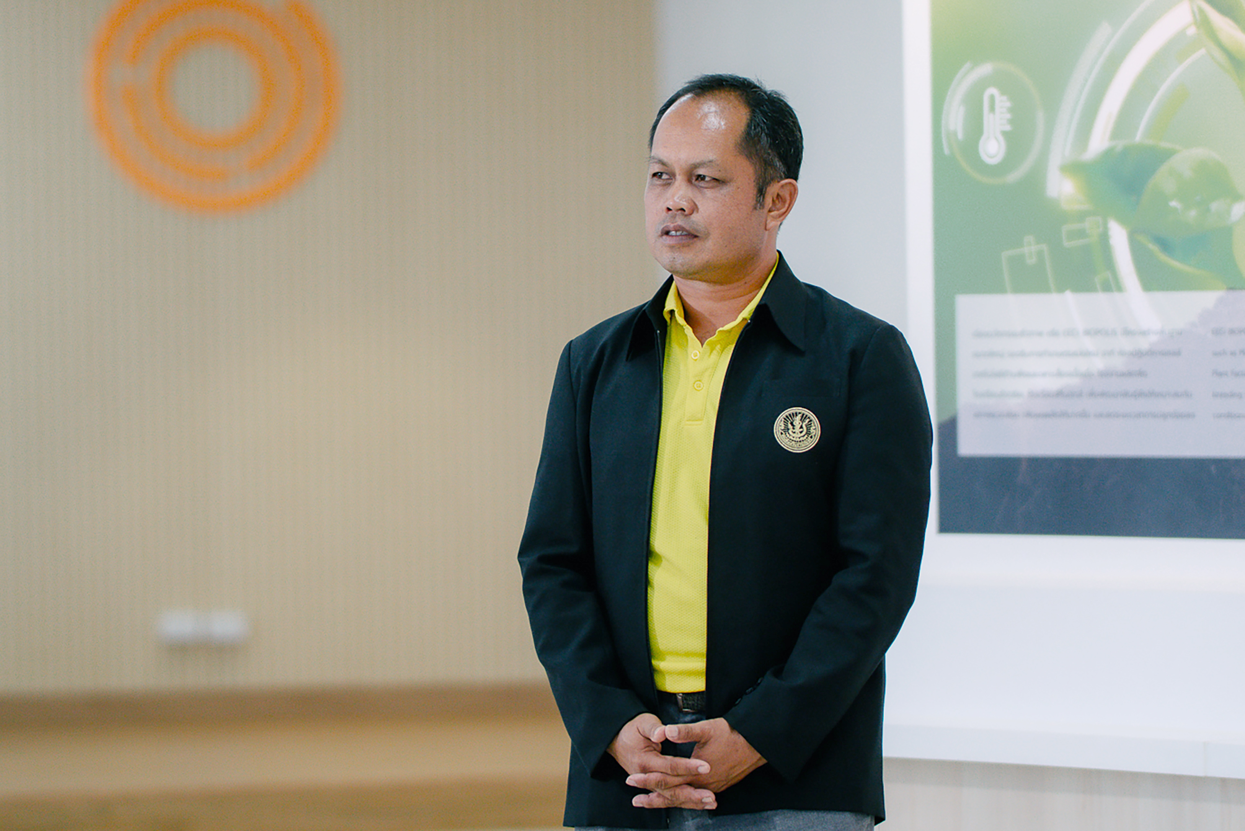 ภาพครึ่งตัว นายวัลลภ ใส่เสื้อแจ็คเก็ตสีดำ ด้านในเป็นเสื้อสีเหลืองอ่อน ทำท่ามือประสานกันอยู่ ด้านหลังเป็นโลโก้ของ EECi 