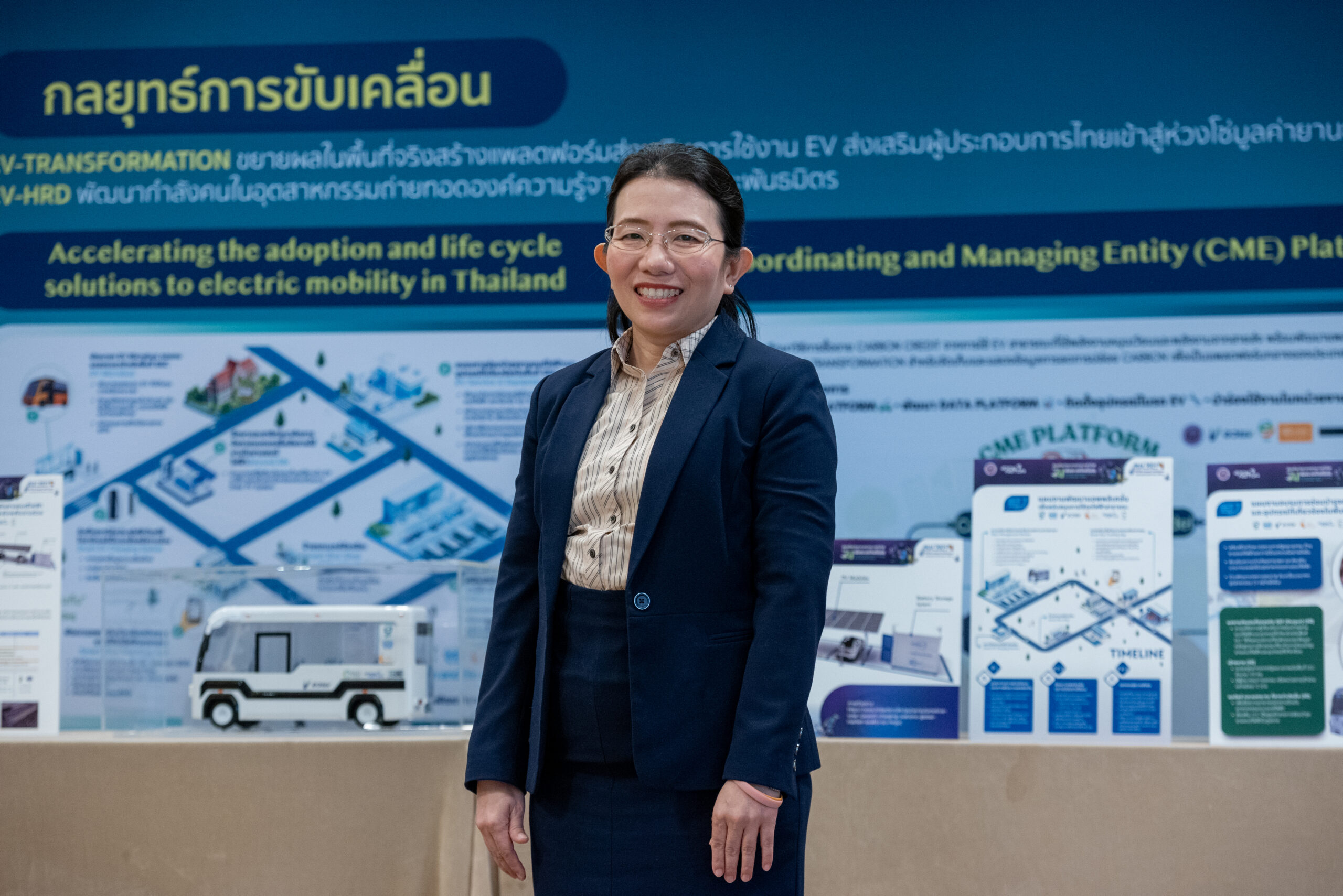 ภาพครึ่งตัวของ ดร.สุมิตรา สวมแว่นสายตา ใส่เสื้อเชิ้ตลายทับด้วยสูทสีกรมท่า ใบหน้ายิ้มแย้ม ยืนอยู่ด้านหน้าบูธนิทรรศการพลังงาน EV 
