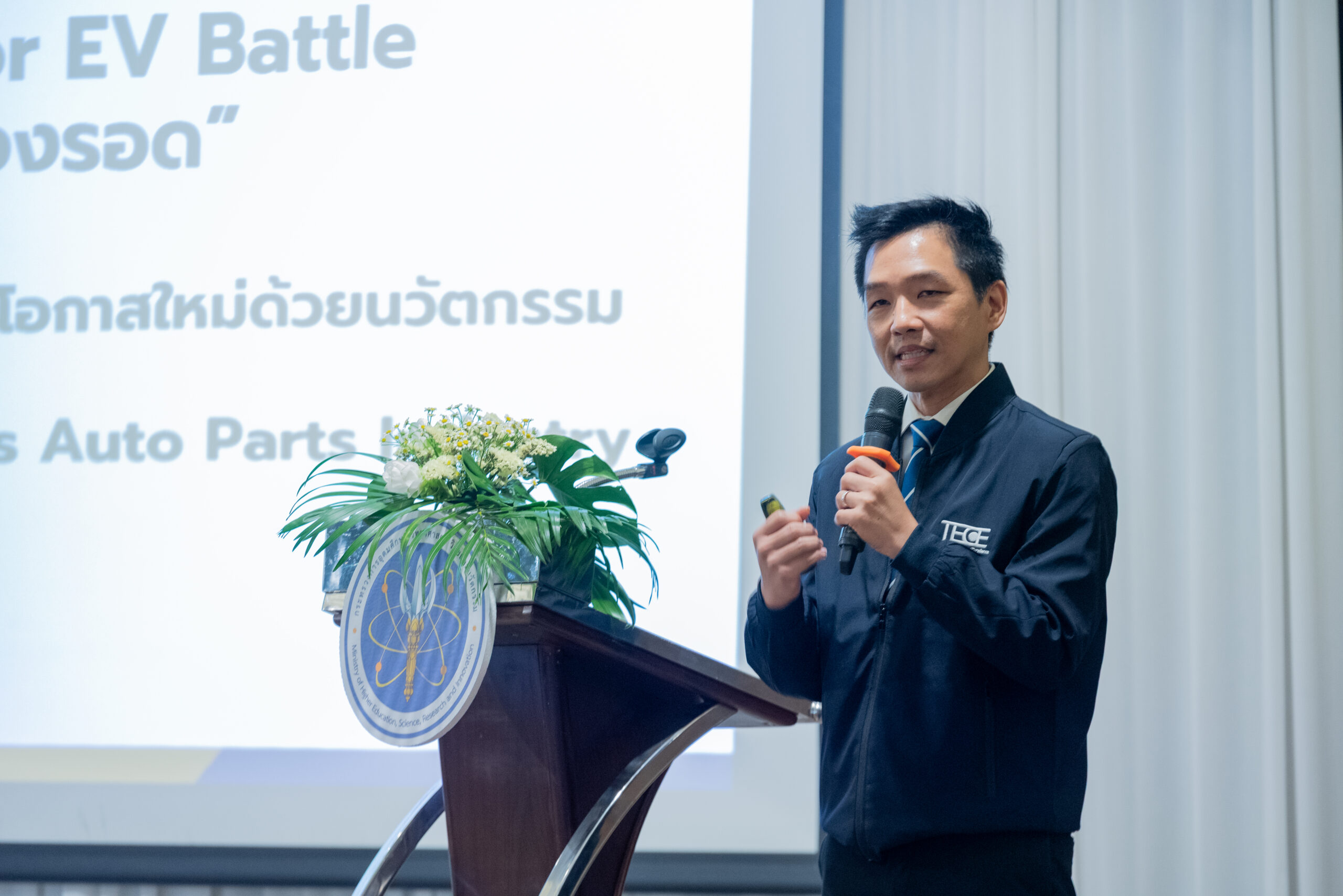 ภาพ ดร.ศราวุธ กำลังจับไมค์พูดข้างๆโพเดียมที่มีดอกไม้ประดับและโโก้ อว. โดยคุณสุพจน์ ใส่เสื้อเชิ้ตขาวผูกไทลายน้ำเงินฟ้า สวมแจ็คเก็ตสีกรมท่า โลโก้ TECE ตรงหน้าซ้าย มีใบหน้ายิ้มแย้ม