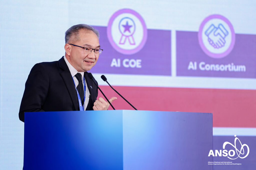 ภาพครึ่งตัวของ ศาสตราจารย์ ดร.ชูกิจ ยืนตรงโพเดี่ยม กำลังพูดด้วยใบหน้ายิ้มแย้ม ด้านหลังเป็นสไลด์นำเสนอ