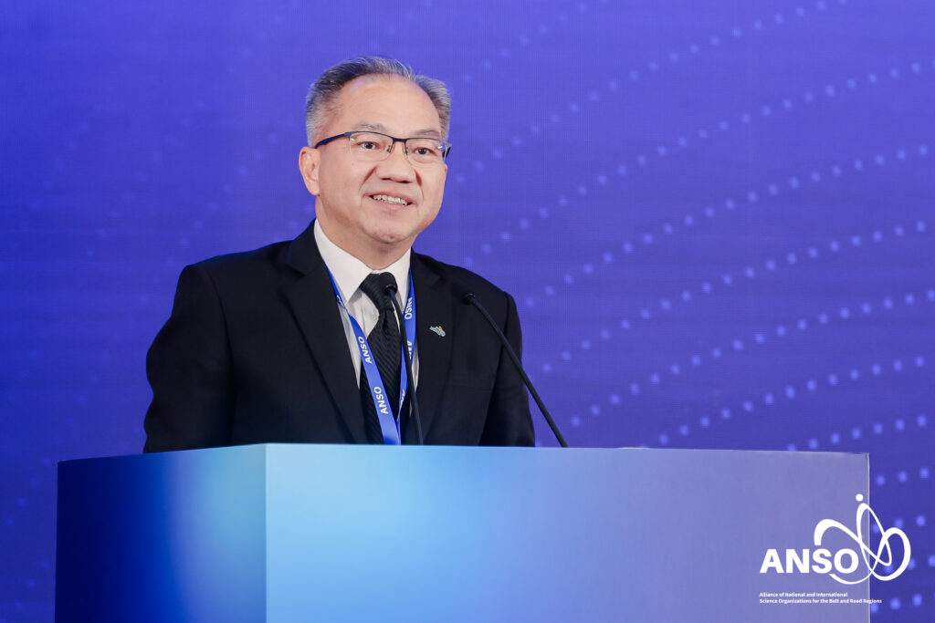 ภาพครึ่งตัวของ ศาสตราจารย์ ดร.ชูกิจ ยืนตรงโพเดี่ยม หน้า Backdrop งาน ANSO ธีมสีน้ำเงิน กำลังพูดด้วยใบหน้ายิ้มแย้ม