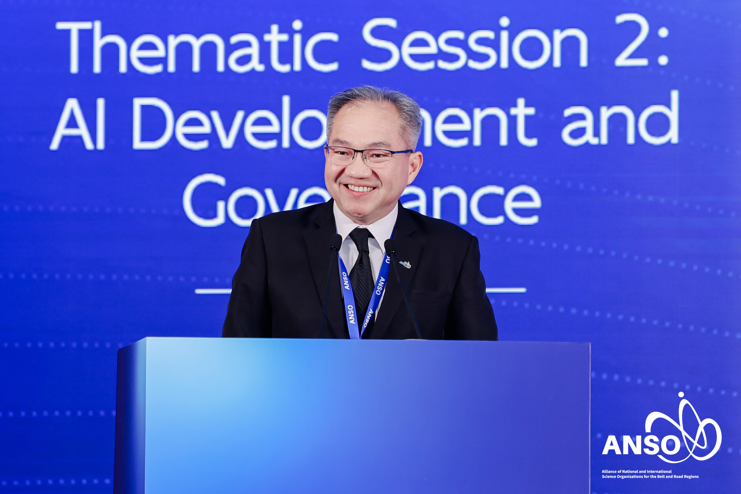 ภาพครึ่งตัวของ ศาสตราจารย์ ดร.ชูกิจ ยืนตรงโพเดี่ยม หน้า Backdrop งาน ANSO ธีมสีน้ำเงิน กำลังพูดด้วยใบหน้ายิ้มแย้ม