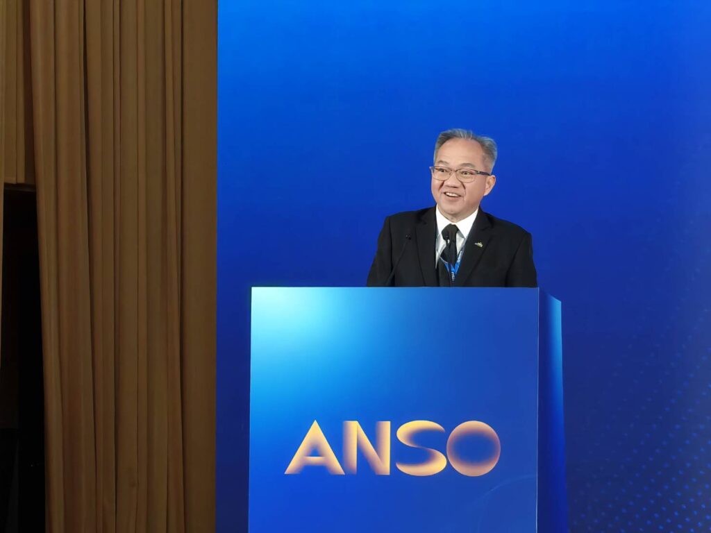 ภาพครึ่งตัวของ ศาสตราจารย์ ดร.ชูกิจ ยืนตรงโพเดี่ยม หน้า Backdrop งาน ANSO ธีมสีน้ำเงิน กำลังพูดด้วยใบหน้ายิ้มแย้ม