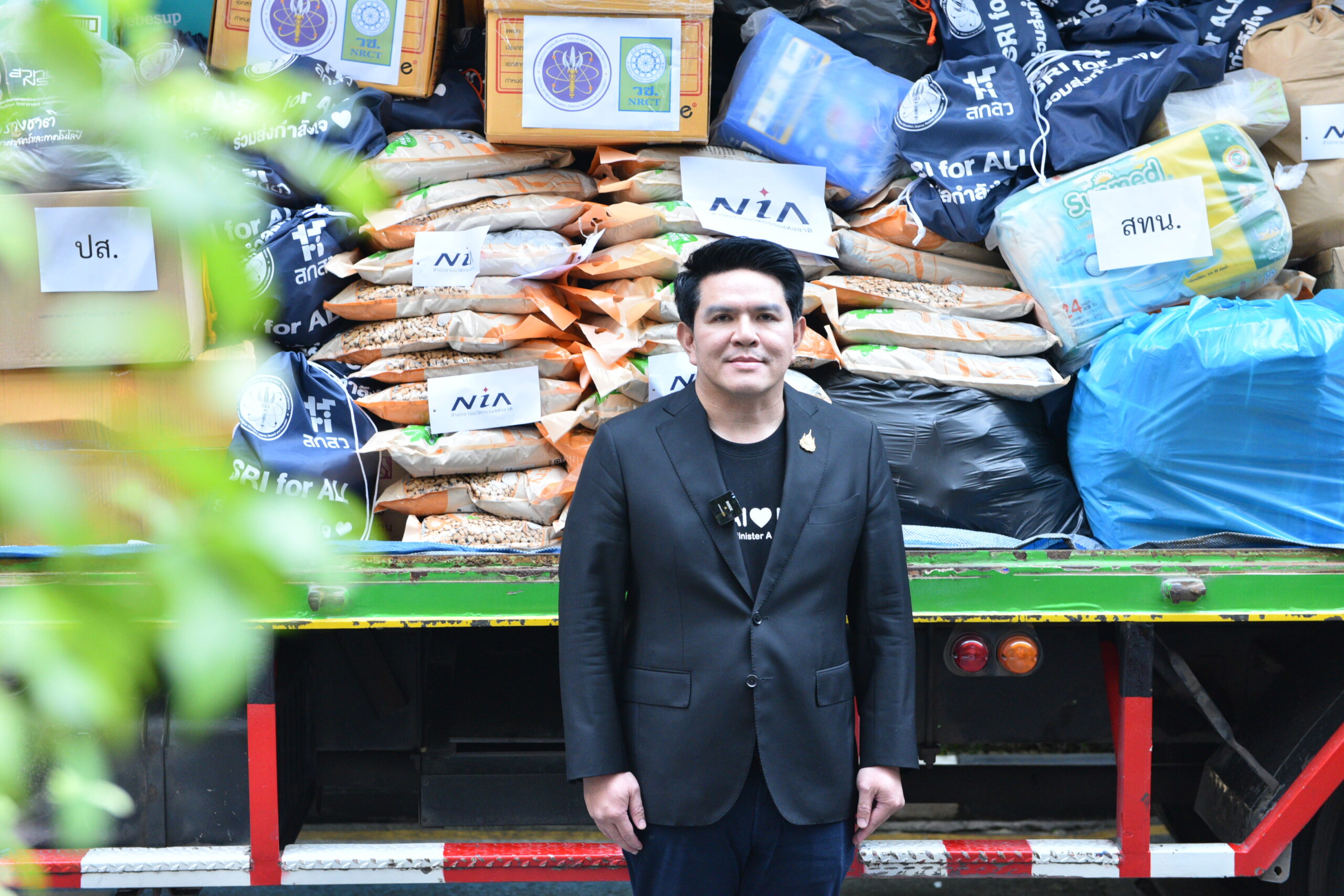 ภาพรัฐมนตรีและคณะทำงานยืนหน้ากองสิ่งของบริจาคที่จัดเรียงอยู่บนรถบรรทุกเตรียมลำเลียงช่วยเหลือผู้ประสบภัยน้ำท่วม