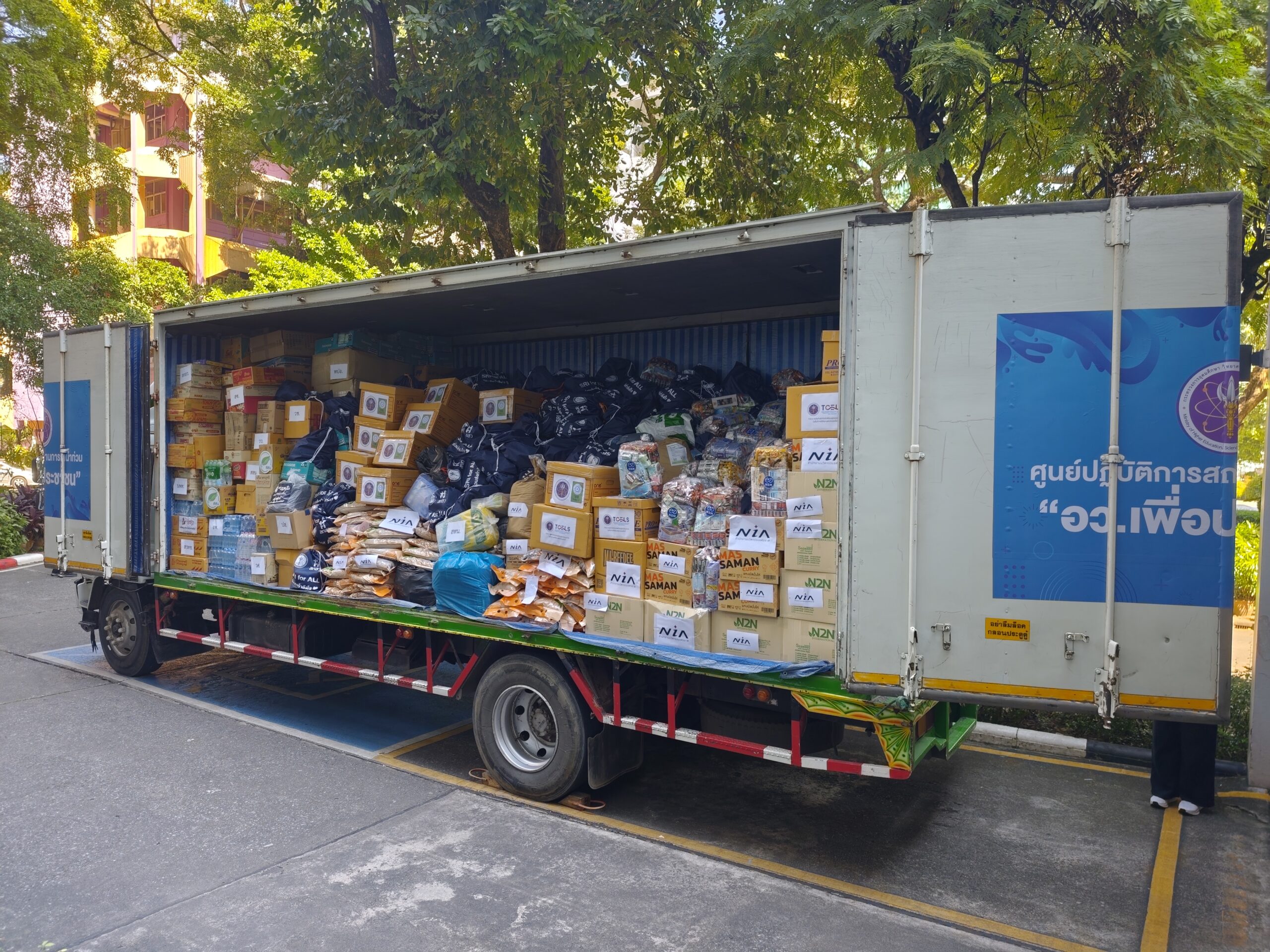 ภาพรัฐมนตรีกระทรวง อว. ยืนหน้ารถบรรทุกสิ่งของบริจาคที่บรรจุข้าวสาร อาหารแห้ง และของใช้จำเป็นจำนวนมาก