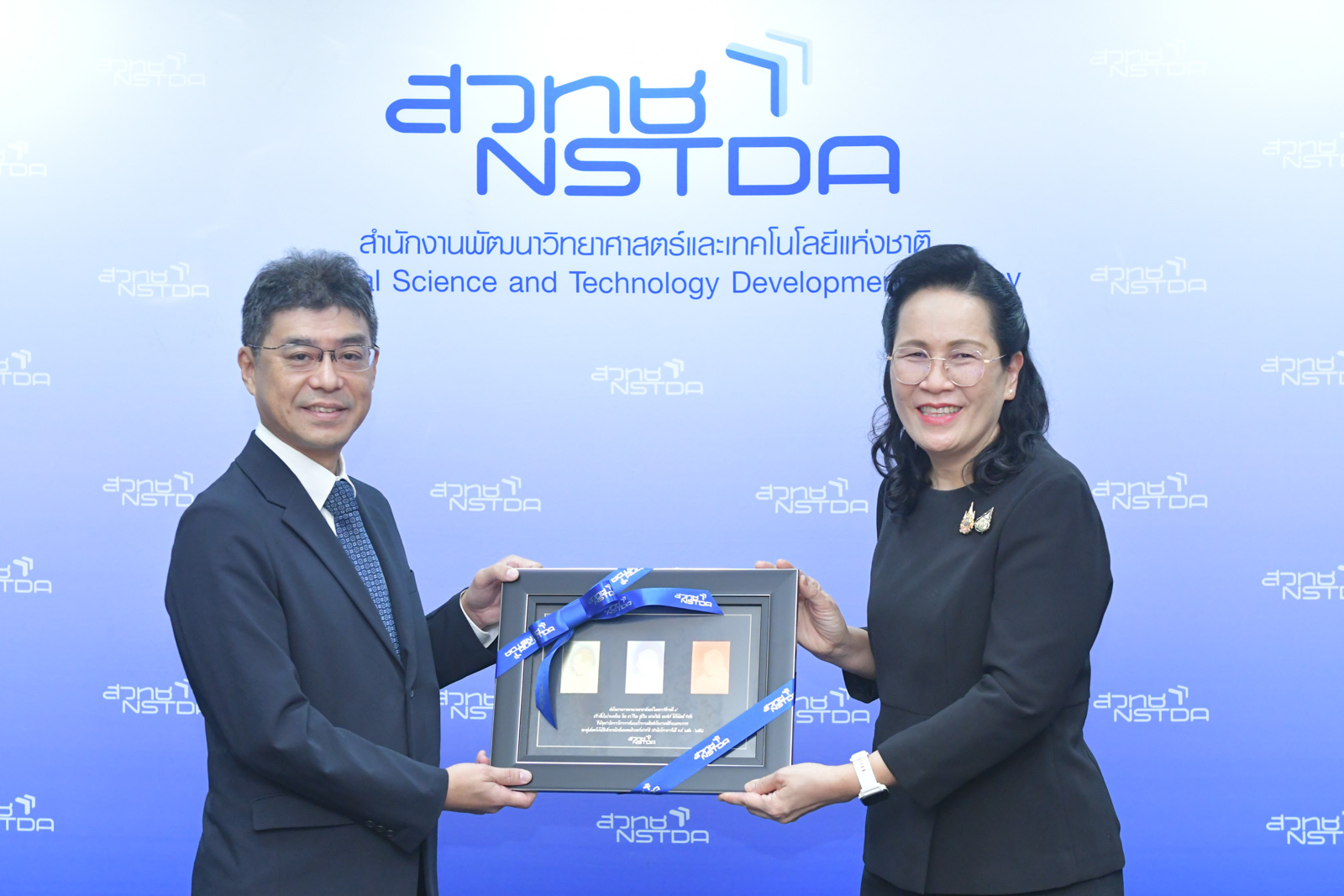 ภาพผู้แทนบริษัทเอกชนมอบของที่ระลึกให้ผู้แทน สวทช. ในพิธีส่งมอบเงินสนับสนุน ณ ฉากหลังโลโก้ NSTDA