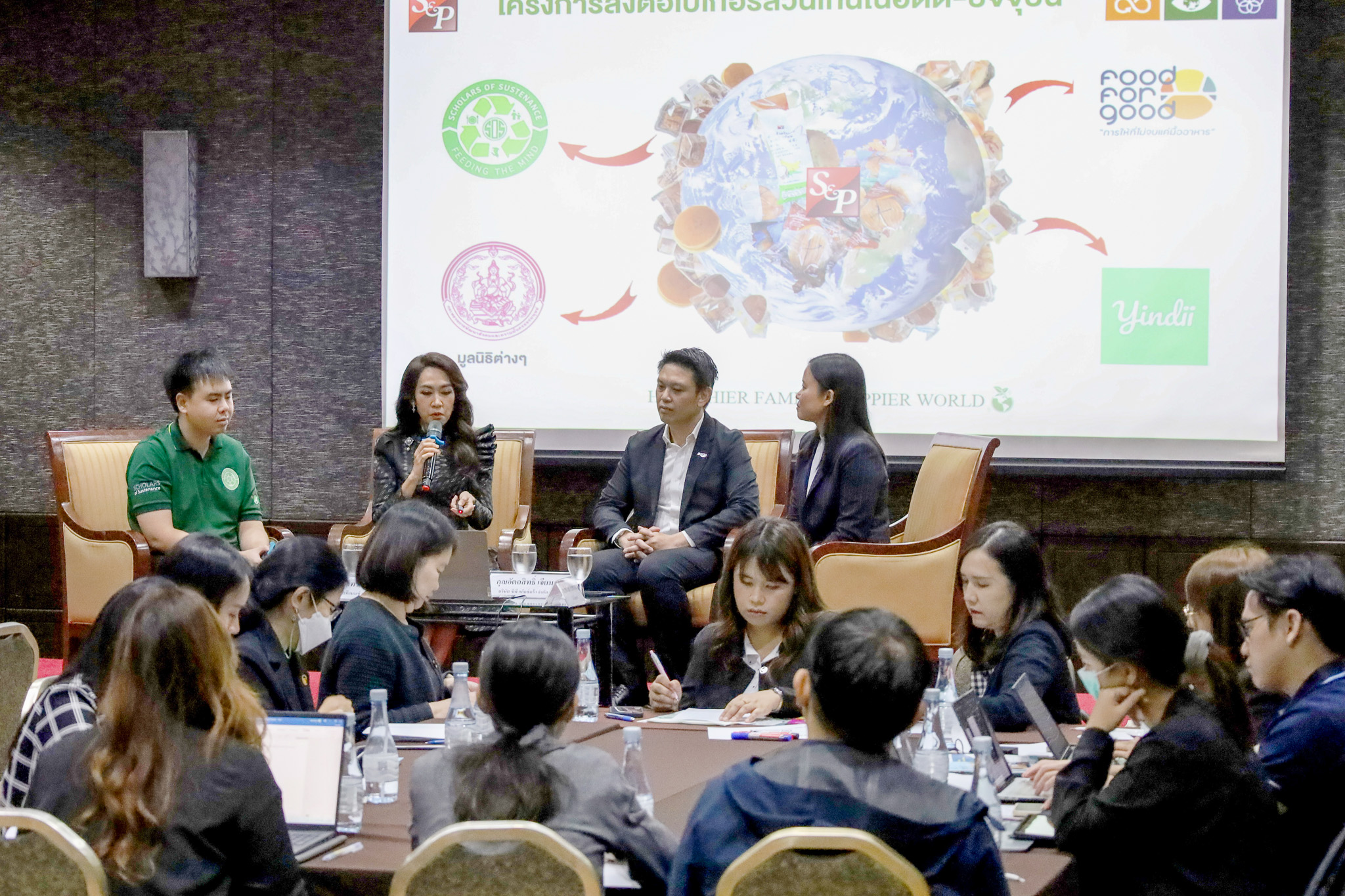 ภาพผู้ร่วมเสวนาสี่คนบนเวที โดยมีสไลด์นำเสนอขนาดใหญ่ด้านหลัง และผู้เข้าร่วมประชุมนั่งรับฟังอยู่ด้านหน้า