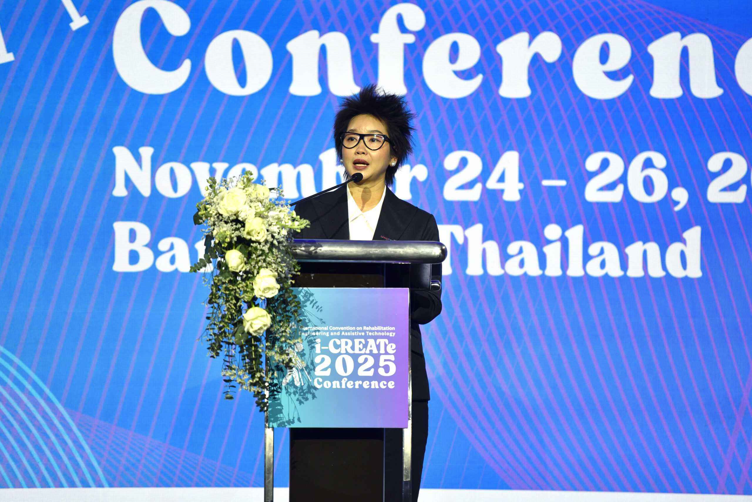 ภาพ ดร.อุรชา กล่าวที่โพเดียม บนเวที ด้านหลังเป็น LED Backdrop งาน ธีมฟ้าม่วง