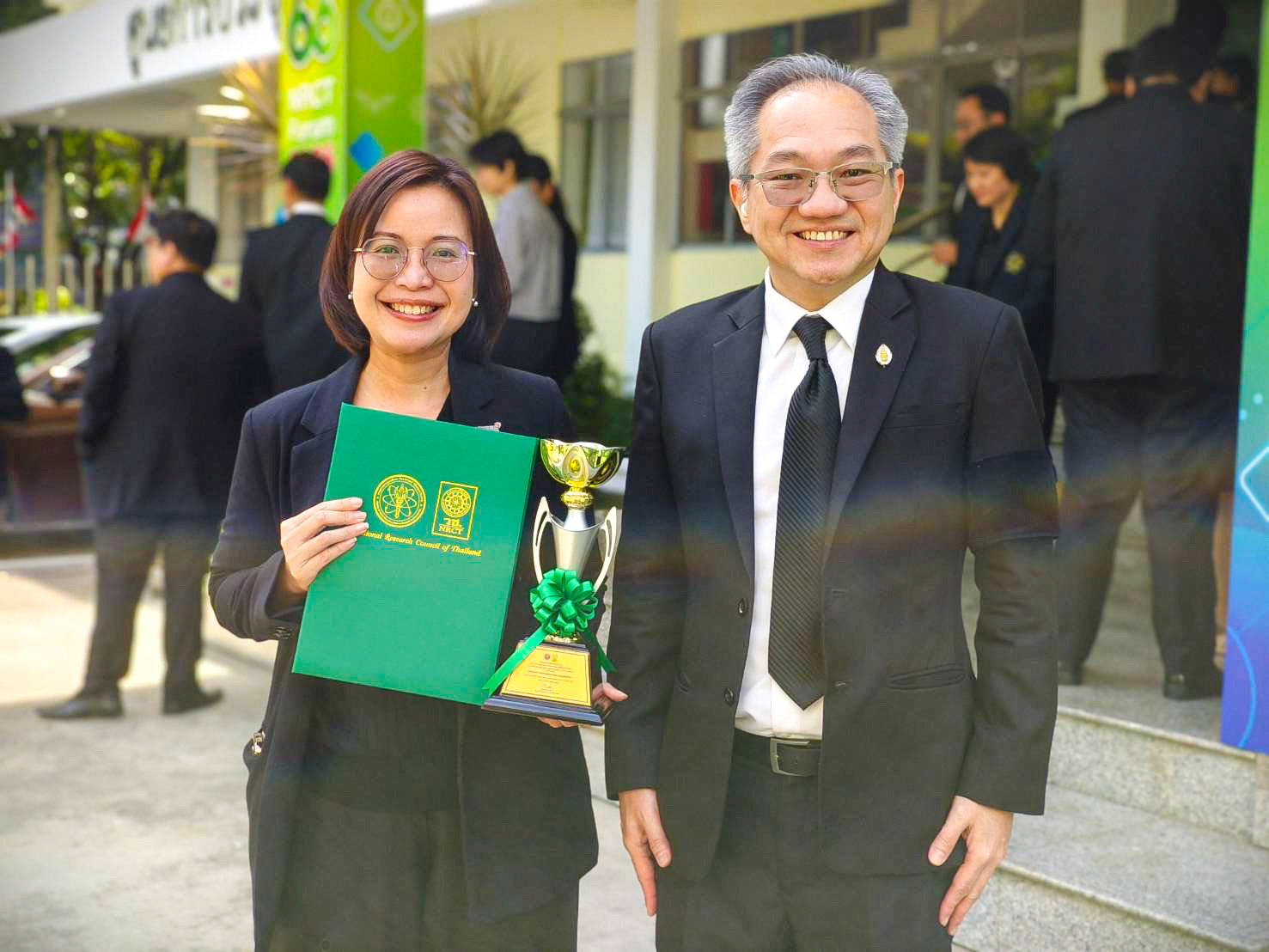 ภาพนักวิจัยร่วมถือถ้วยรางวัลและเอกสารรางวัล NRCT AWARD ถ่ายภาพร่วมกับผู้บริหารบริเวณด้านหน้าอาคารสถานที่จัดงาน