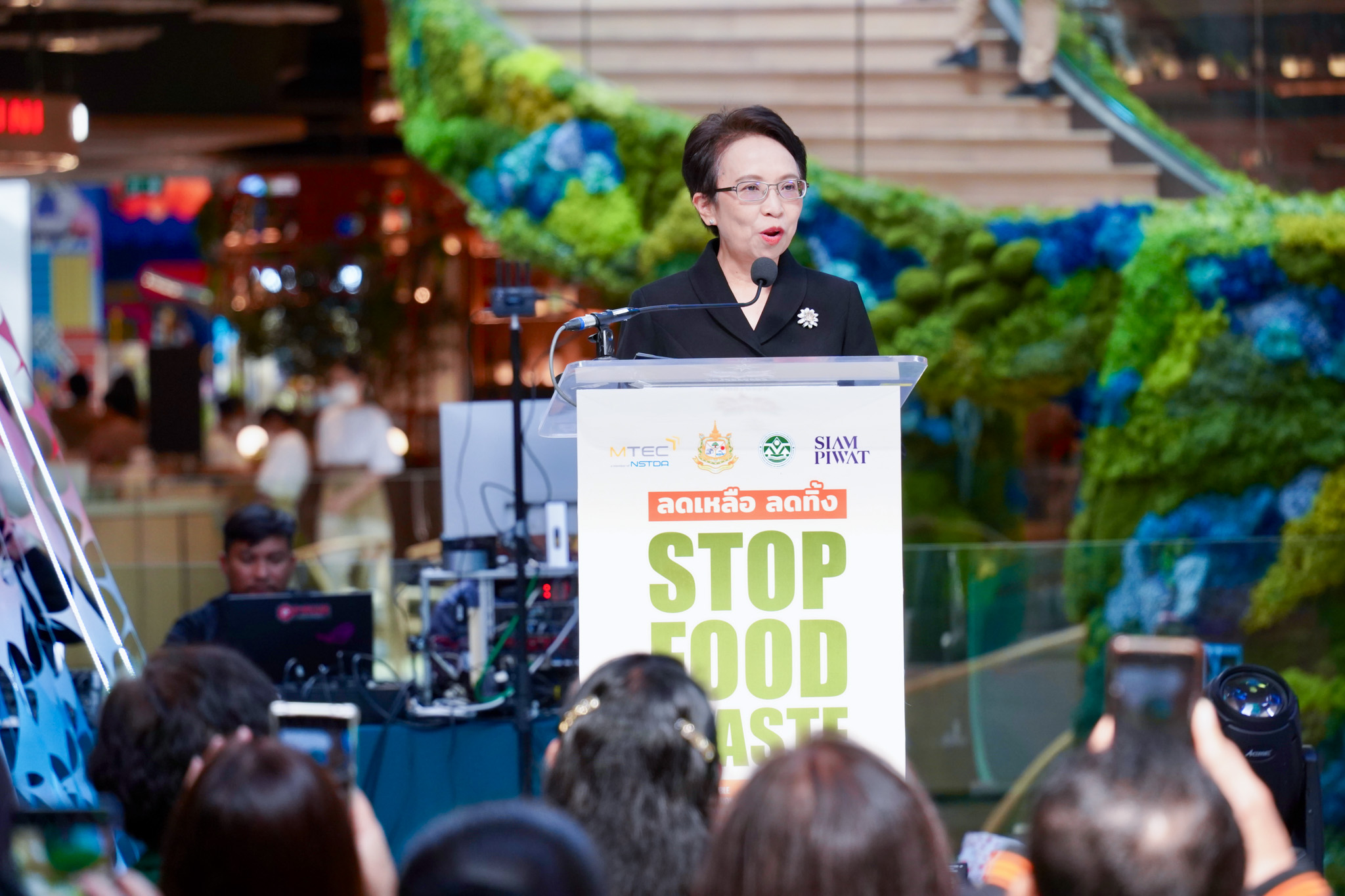 ภาพ นางสาวปรีญาพร ยืนกล่าวที่โพเดียมบนเวที ในงานพิธีเปิดโครงการ ลดเหลือ ลดทิ้ง Stop Food Waste SDG 12.3
