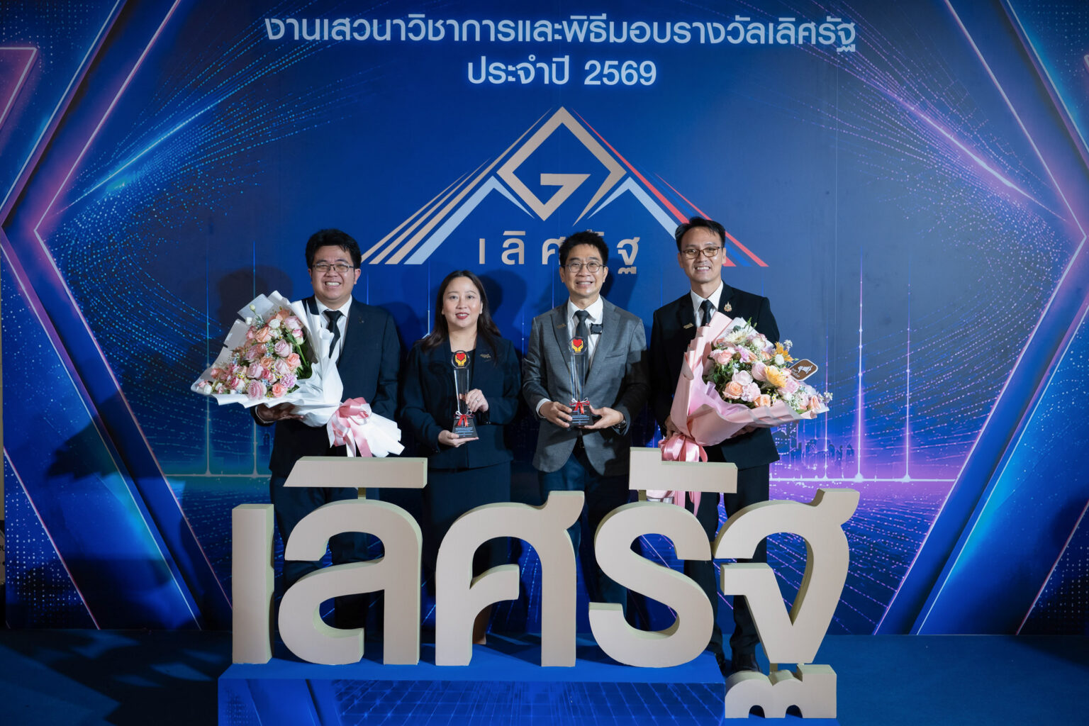 ผู้บริหาร สวทช. 4 ท่าน ถือโล่รางวัลเลิศรัฐ ประจำปี 2569 และช่อดอกไม้ หน้าป้ายเลิศรัฐ ณ เมืองทอ